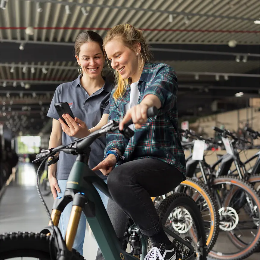 Zwei Personen stehen in einem Fahrradgeschäft. Eine Person sitzt auf einem grünen E-Bike und trägt eine karierte Jacke sowie schwarze Jeans und schwarze Schuhe. Die andere Person steht daneben und hält ein Smartphone. Im Hintergrund sind mehrere Fahrräder und eine moderne Einrichtung zu sehen.