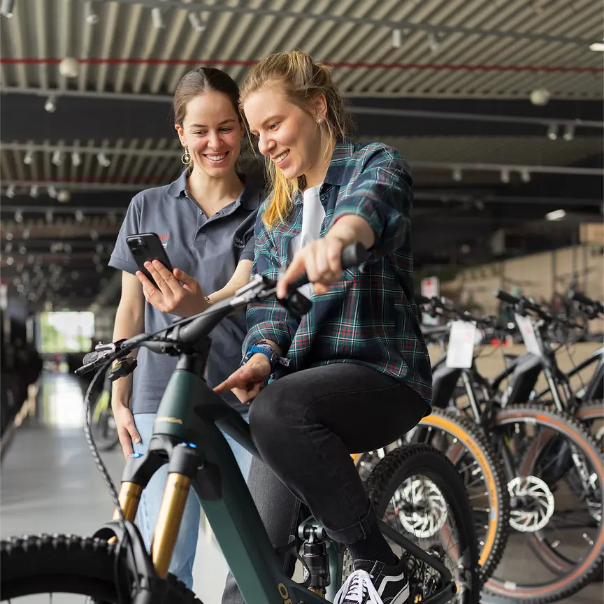 Zwei Personen stehen in einem Fahrradgeschäft. Eine Person sitzt auf einem grünen E-Bike und trägt eine karierte Jacke sowie schwarze Jeans und schwarze Schuhe. Die andere Person steht daneben und hält ein Smartphone. Im Hintergrund sind mehrere Fahrräder und eine moderne Einrichtung zu sehen.