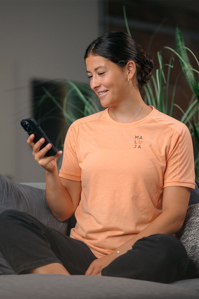 Una donna con un top arancione è seduta a gambe incrociate su un divano grigio e guarda il suo Smartphone. Sullo sfondo si può vedere una pianta.