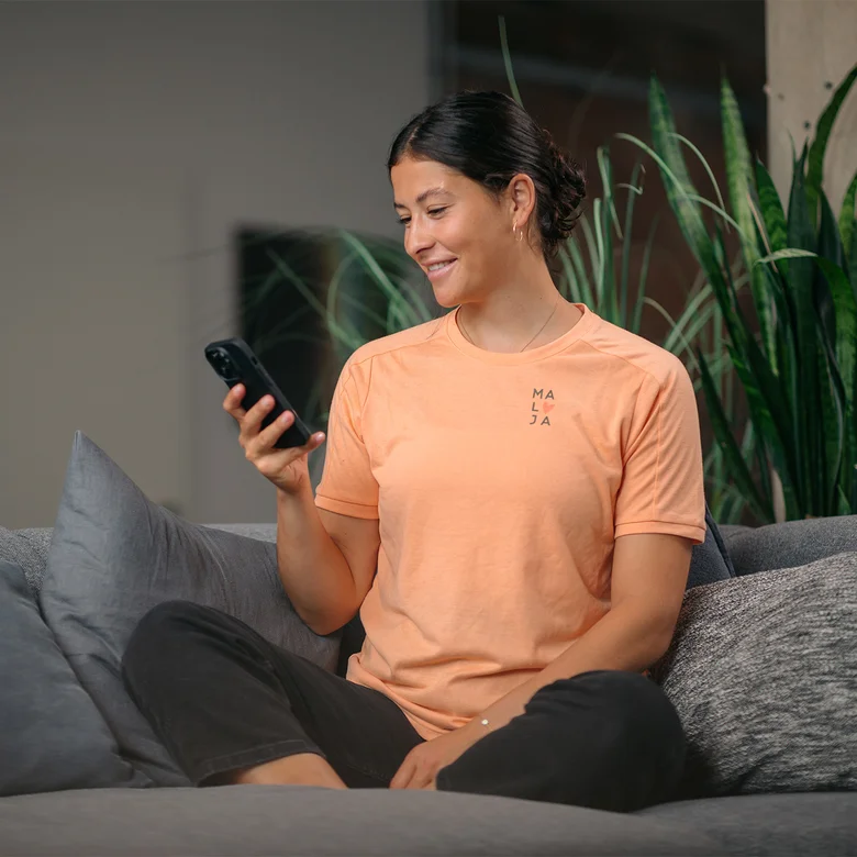 A woman in an orange top sits cross-legged on a gray sofa and looks at her smartphone. In the background, you can see a plant.