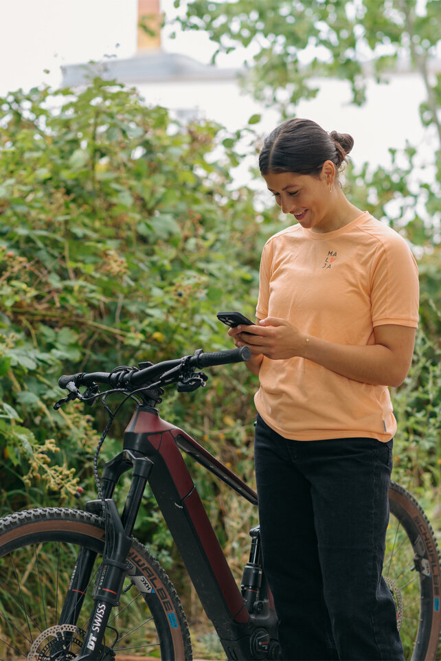 A woman stands next to her eBike, holding her smartphone and smiling.