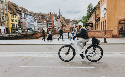 Eine Person fährt ein weißes E-Bike mit Bosch Komponenten auf einer Straße in einer Stadt mit historischen Gebäuden. Im Hintergrund sind bunte Häuser und eine Kirche mit einem hohen Turm zu sehen. Mehrere Fußgänger gehen auf dem Gehweg entlang eines Kanals.