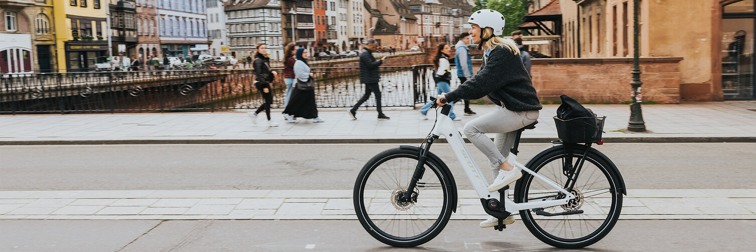 Eine Person fährt ein weißes E-Bike mit Bosch Komponenten auf einer Straße in einer Stadt mit historischen Gebäuden. Im Hintergrund sind bunte Häuser und eine Kirche mit einem hohen Turm zu sehen. Mehrere Fußgänger gehen auf dem Gehweg entlang eines Kanals.