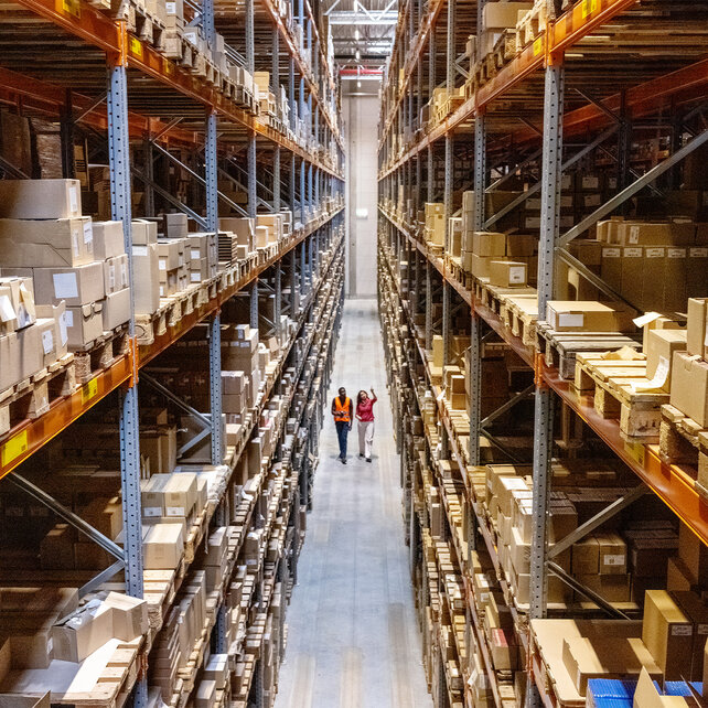 Shot of a warehouse with high shelves and packages, in the distance you can see two people long walking a path between two shelves.