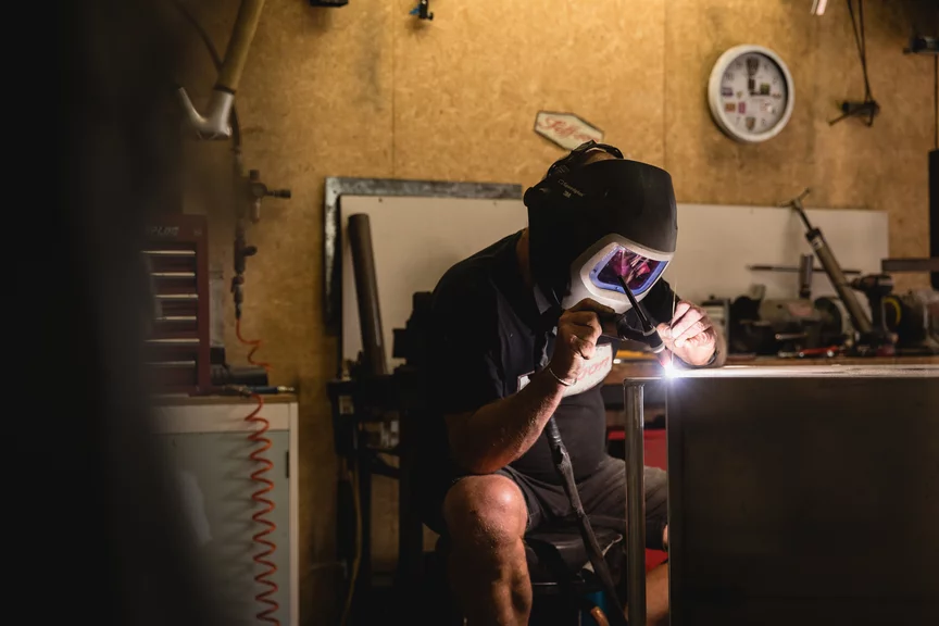 A man wearing protective equipment and a welding helmet is welding at a table in a workshop.