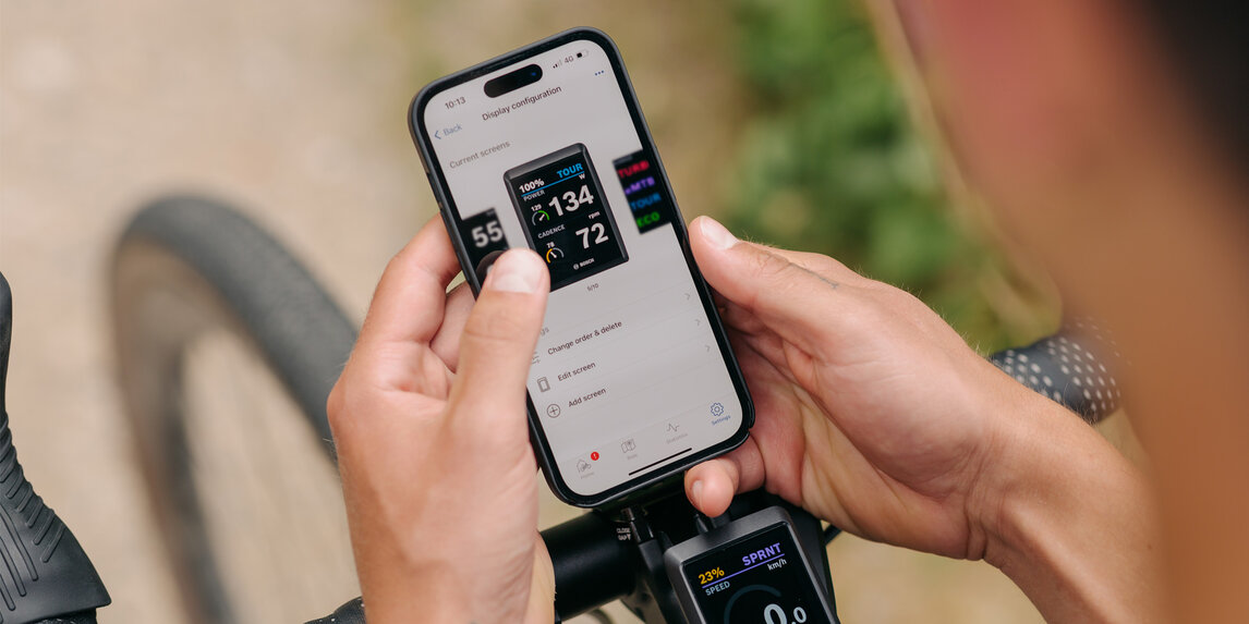 A person holds their Smartphone in hand, with the eBike Flow App open. In the background, you can see the handlebar and front wheel of an eBike.
