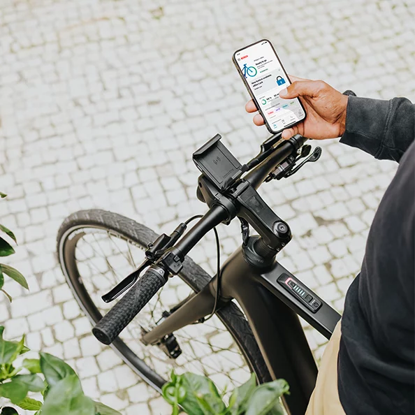 En person står på en el-sykkel og holder en smarttelefon i hånden, der eBike Flow-appen er åpen.