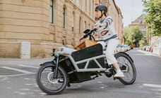 The Cargo eBike rider drives down a long road, in the background you can see a building.