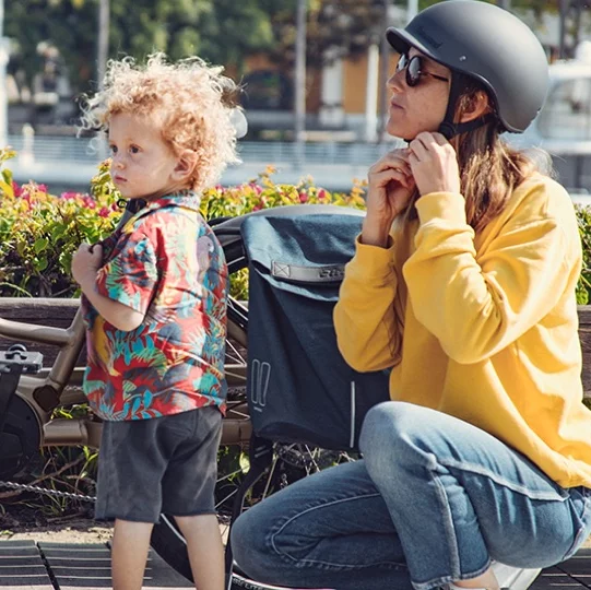 Frau kniet vor ihrem eBike und schnallt ihren Fahrradhelm zu, ihr Kind steht vor ihr.