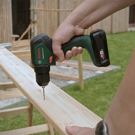 Una persona avvita una vite su una tavola di legno all'aperto utilizzando un trapano verde Bosch batteria.