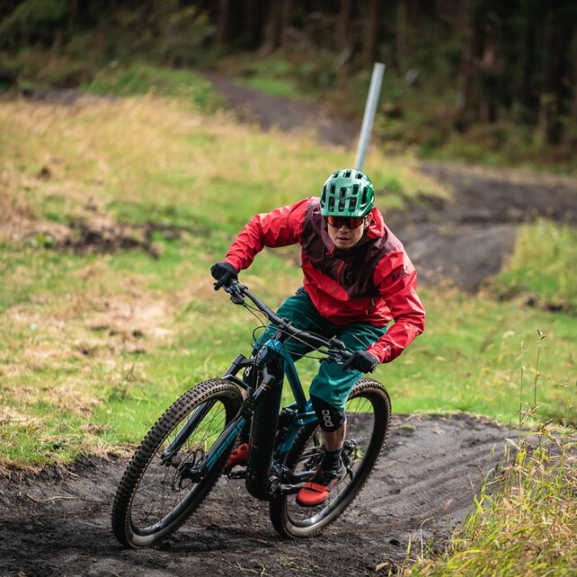 A person rides an e-mountain bike on a muddy path in a green, wooded environment. She is wearing a green helmet, a red jacket, and green pants.