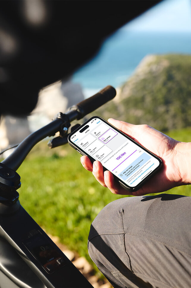 Une personne est assise sur son VAE noir et tient un smartphone dans sa main. En arrière-plan, on distingue une mer floue, l’horizon, des rochers et une prairie. L’écran du smartphone affiche les réglages des modes de conduite dans l'application eBike Flow.