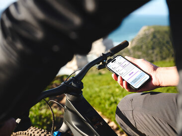 Une personne est assise sur son VAE noir et tient un smartphone dans sa main. En arrière-plan, on distingue une mer floue, l’horizon, des rochers et une prairie. L’écran du smartphone affiche les réglages des modes de conduite dans l'application eBike Flow.