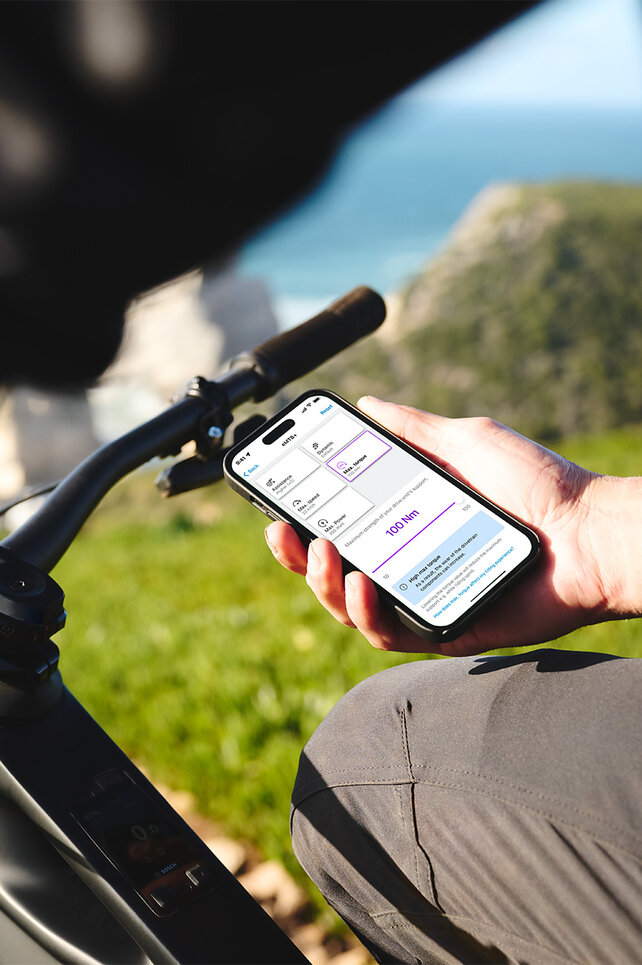 Eén persoon zit op zijn zwarte eBike en houdt een smartphone in zijn hand. Op de achtergrond zijn een wazige zee, de horizon, rotsen en een weide te zien. Het display van de smartphone toont de instellingen voor de rijmodi in de eBike Flow-app.