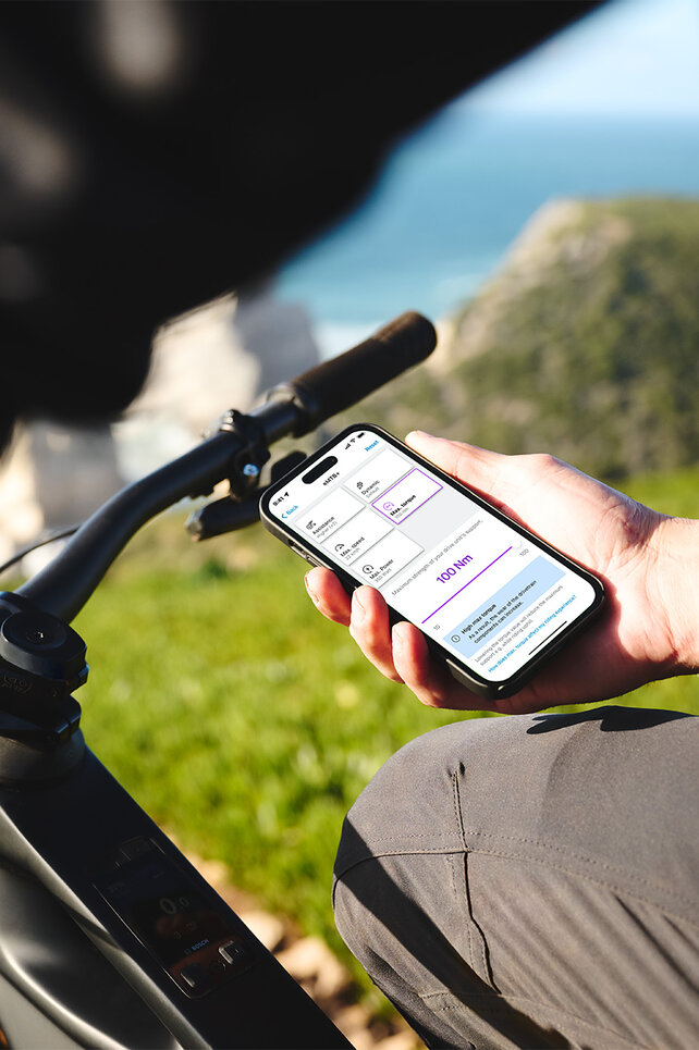 Osoba sedí na černém eBike a v ruce drží telefon. V pozadí je rozmazané moře, obzor, skály a louka. Displej telefonu zobrazuje nastavení jízdních režimů v aplikaci eBike Flow.