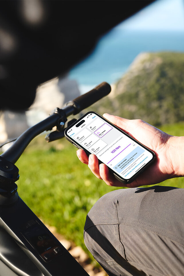 En person sitter på en sort elsykkel og holder en telefon i hånden. I bakgrunnen skimtes et uskarpt hav, horisonten, steiner og en eng. Telefonskjermen viser innstillinger for kjøremodusene i eBike Flow-appen.