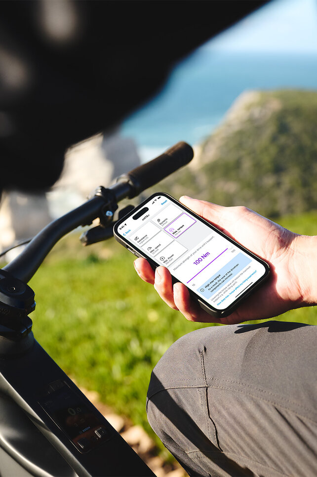 Une personne est assise sur son VAE noir et tient un smartphone dans sa main. En arrière-plan, on distingue une mer floue, l’horizon, des rochers et une prairie. L’écran du smartphone affiche les réglages des modes de conduite dans l'application eBike Flow.