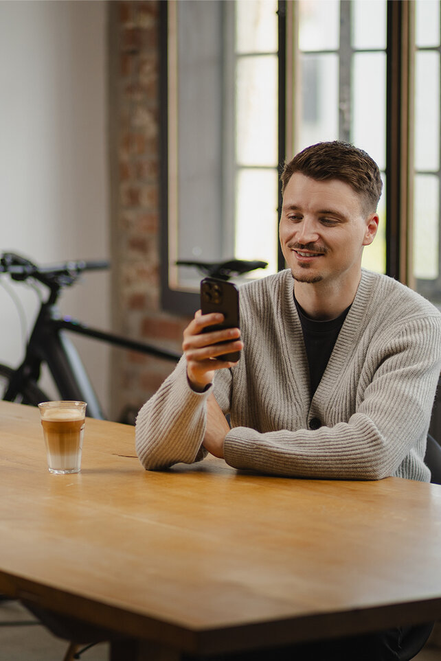 A young man sits at a wooden table, smiling as he looks at his smartphone. On the table, there is a glass of latte macchiato and a small plant. An eBike is visible in the background.
