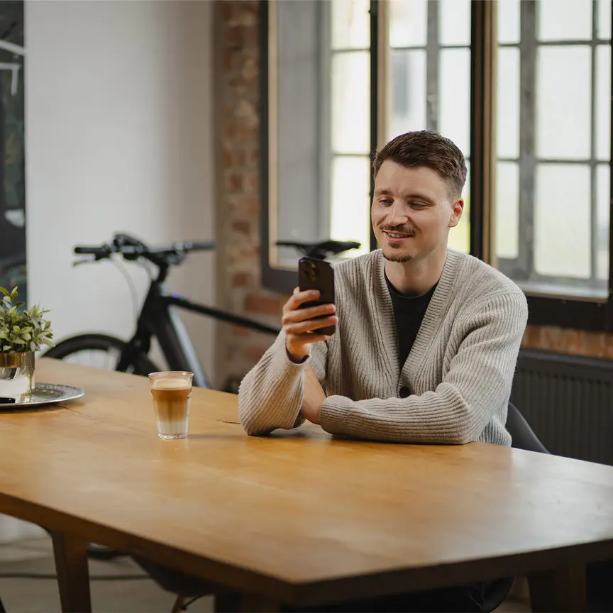 Ein junger Mann sitzt lächelnd an einem Holztisch und blickt auf sein Smartphone. Auf dem Tisch stehen ein Glas Latte Macchiato und eine kleine Pflanze. Im Hintergrund ist ein eBike zu sehen.