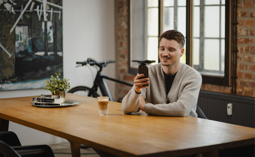 Ein junger Mann sitzt lächelnd an einem Holztisch und blickt auf sein Smartphone. Auf dem Tisch stehen ein Glas Latte Macchiato und eine kleine Pflanze. Im Hintergrund ist ein eBike zu sehen.