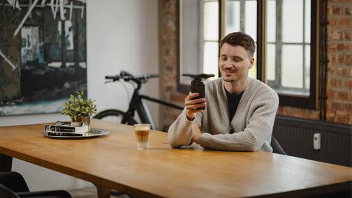 A young man sits at a wooden table, smiling as he looks at his smartphone. On the table, there is a glass of latte macchiato and a small plant. An eBike is visible in the background.