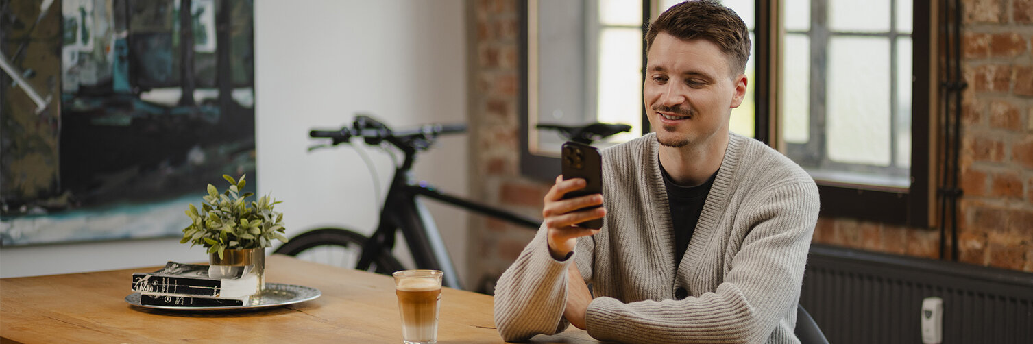 Un jeune homme est assis en souriant à une table en bois et regarde son smartphone. Sur la table, un verre de latte macchiato et une petite plante. Un VAE est visible en arrière-plan.