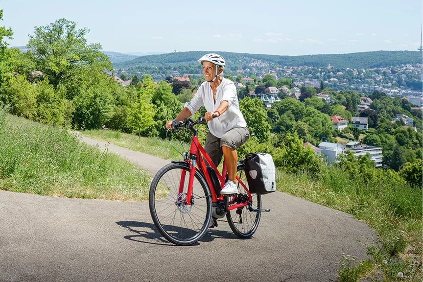 Une femme eBike utilisateur souriante monte un chemin escarpé et sinueux sur un eBike rouge. À côté du chemin, vous pouvez voir des herbes, des buissons et des arbres, en arrière-plan, vous pouvez voir un paysage avec des maisons et des montagnes verts.