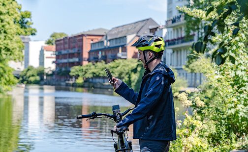 Ein Mann steht mit seinem eBike an einem Fluss und schaut auf ein Smartphone.