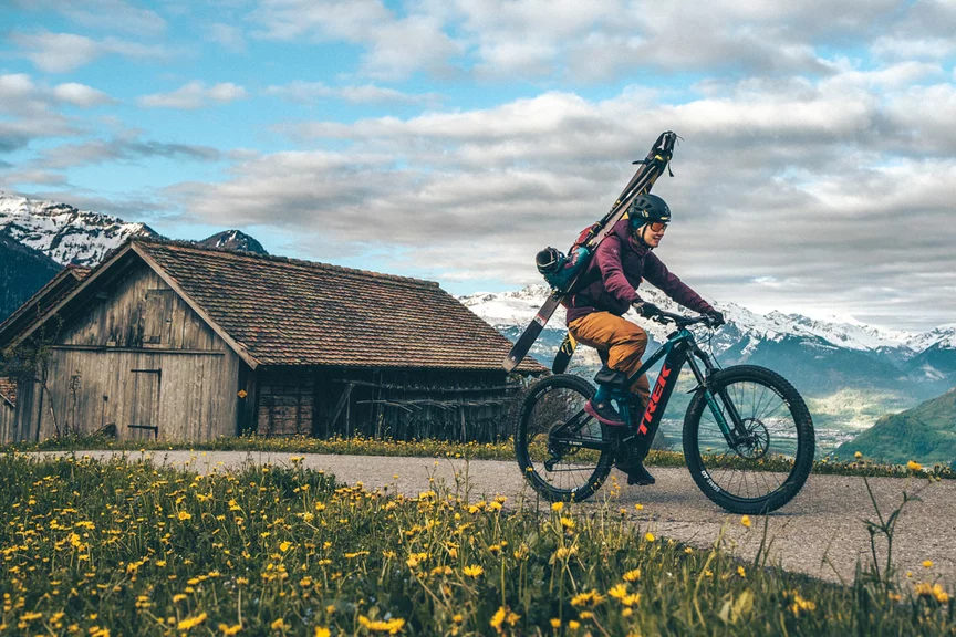 Une cycliste VAE roule sur un chemin de terre avec des skis attachés à son sac à dos. À l’arrière-plan, on aperçoit une cabane, la nature et les montagnes.