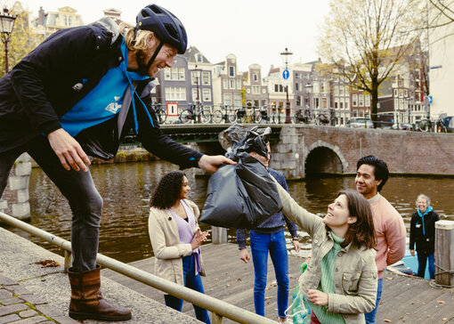 Een vrouw op een steiger geeft een vuilniszak aan een man met een fietshelm. Ze glimlachen allebei.