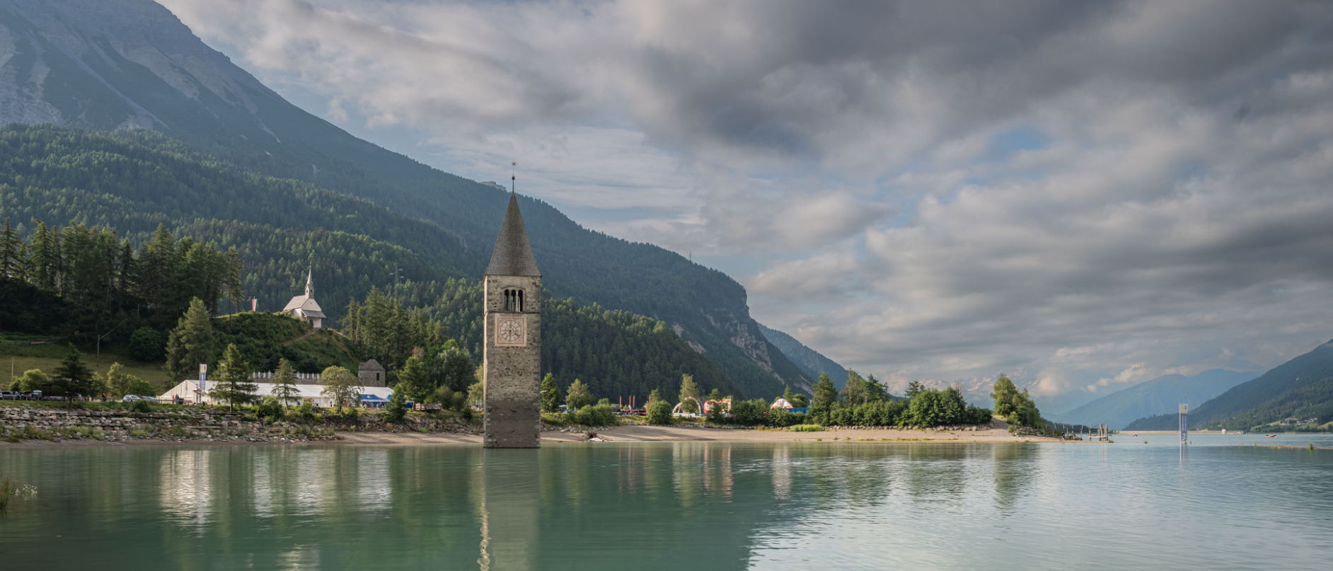 Eine Stadt liegt am Fuße eines Berges direkt an einem See. Ein Kirchturm steht am Ufer, der See ist glatt und blau-grünlich, im Hintergrund erstrecken sich Berge und der Himmel über dem See ist blau und bewölkt. 