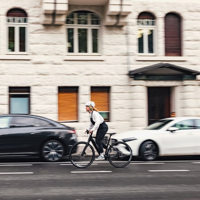 A person rides a black e-bike and is wearing a white helmet and black clothing. In the background, there are parked cars and a building with white stone walls and several windows.
