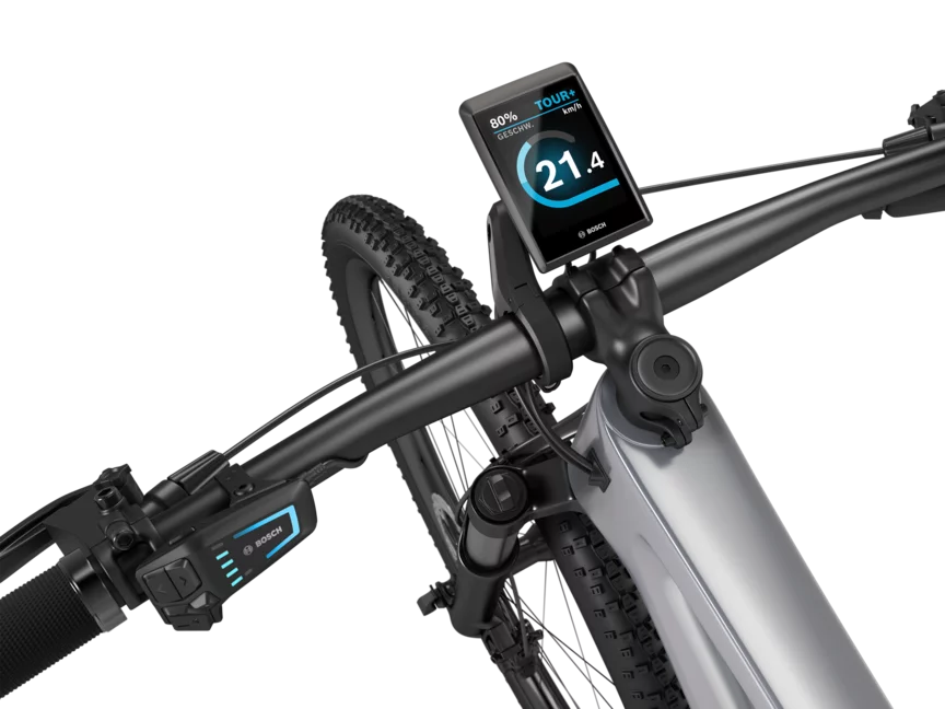 Un VTT à assistance électrique argenté avec un guidon noir, isolé sur fond blanc. Un Kiox 500 est monté sur le guidon. L'écran affiche la vitesse en km/h, le niveau de batterie et le mode de conduite.