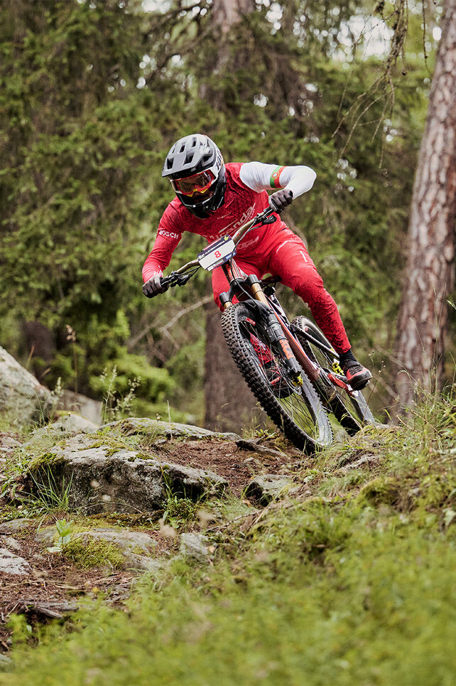 Un VTT à assistance électrique, eMountainbike, Fahrer, vêtu d’un costume rouge descend un chemin forestier au-dessus de pierres.