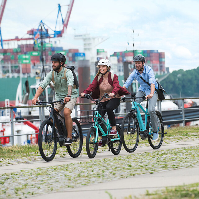 Tres eBike pilotos pasan uno detrás del otro por un puerto, al fondo se pueden ver barcos.