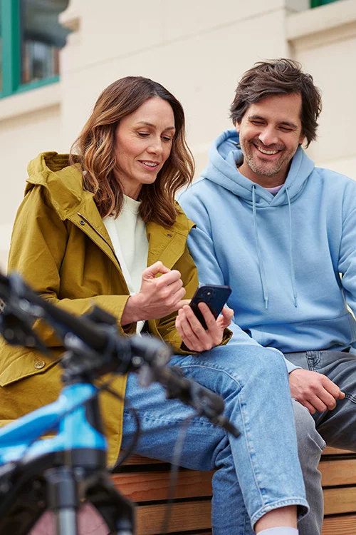 Una mujer y un hombre están sentados juntos riendo mientras miran un teléfono móvil. En primer plano se ve un eBike.