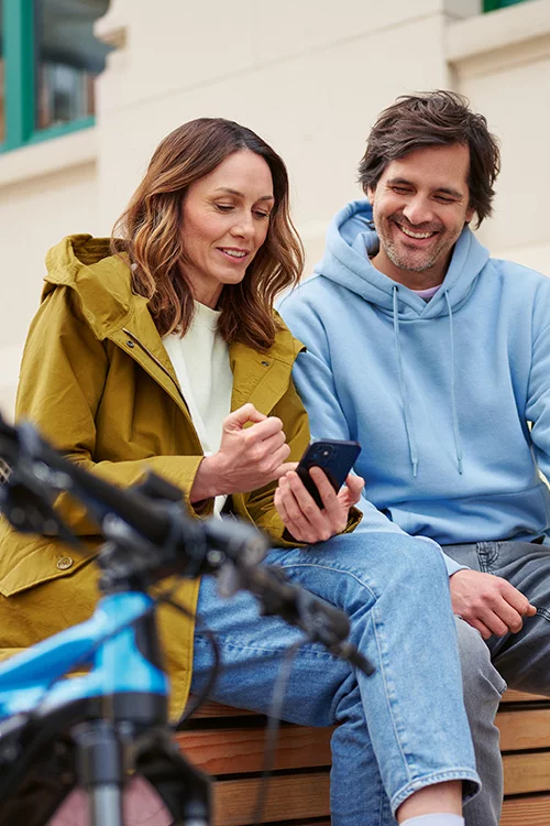 Žena a muž sedí vedle sebe a smějí se, když se dívají na smartphone. V popředí je vidět eBike.