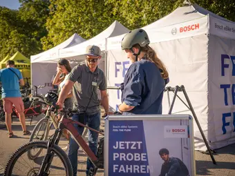 A man in a Bosch polo shirt stands with a woman in front of an eBike in an urban environment. Behind them, there are pavilions with the Bosch logo and more eBikes.