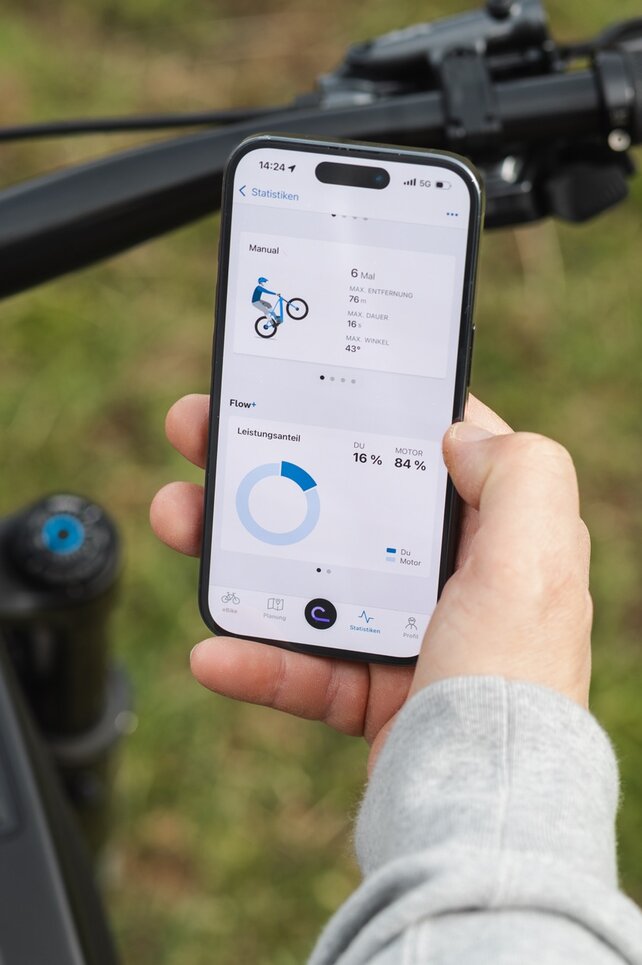 Nahaufnahme einer Hand, die ein Smartphone über einem eBike-Lenker hält. Auf dem Display wird in der eBike Flow App die Trick-Statistik für Manuals und der Leistungsanteil von Fahrer und Motor angezeigt.