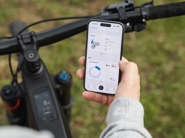 Nahaufnahme einer Hand, die ein Smartphone über einem eBike-Lenker hält. Auf dem Display wird in der eBike Flow App die Trick-Statistik für Manuals und der Leistungsanteil von Fahrer und Motor angezeigt.