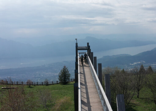 Deux Vététiste traversent un pont en bois. À l’arrière-plan, on aperçoit un paysage montagneux verdoyant et un grand lac.
