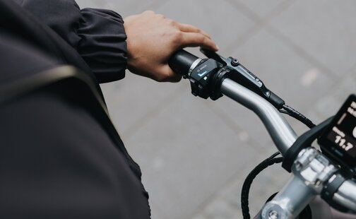 Nærbilde fra førerens perspektiv som viser en hånd på styrehåndtaket på en elsykkel. Tommelen hviler på LED-kontrolleren, der LED indikator lyser. Fokuset er på direkte kontroll og interaksjon med elsykkel teknologi.
