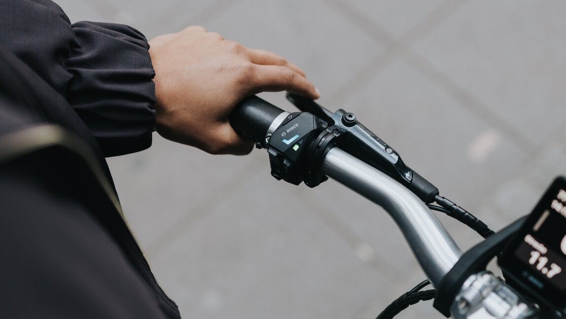 Nærbillede fra rytterens perspektiv, hvor en hånd hviler på styrets greb på en elcykel. Tommelfingeren ligger på LED-controlleren, hvor LED skærm lyser. Fokus er på direkte kontrol og interaktion med eBike teknologi.