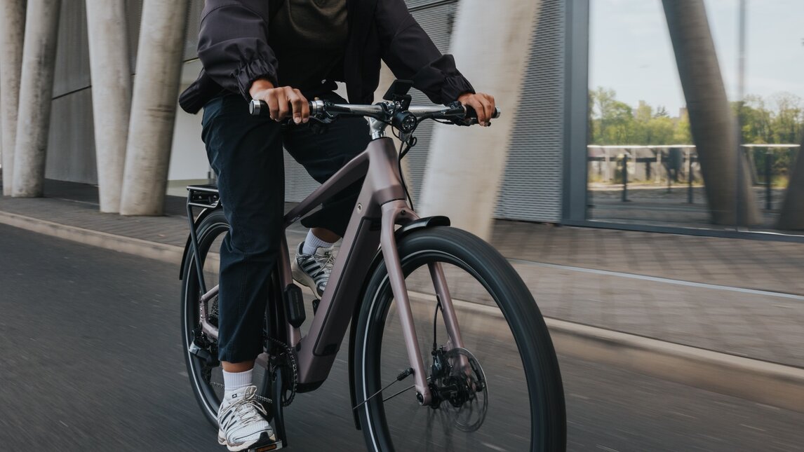 A woman in business-casual attire rides a modern eBike with a helmet past an impressive architectural façade of concrete and glass. The shot captures the dynamics of the urban commute.