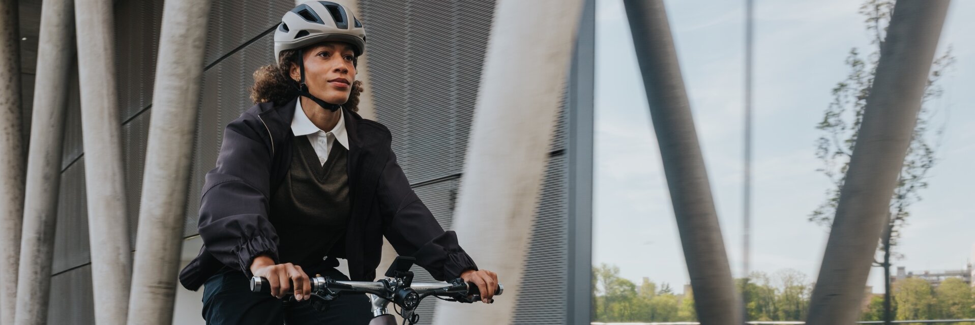 A woman in business-casual attire rides a modern eBike with a helmet past an impressive architectural façade of concrete and glass. The shot captures the dynamics of the urban commute.