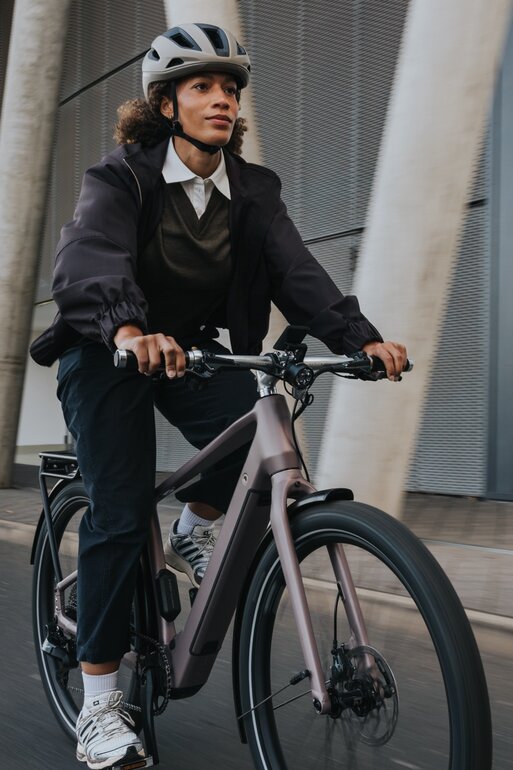 Una donna in stile business-casual, indossando un casco, guida un eBike moderno davanti a una facciata architettonica imponente in cemento e vetro. L'inquadratura mette in risalto la dinamica del percorso urbano per i pendolari.