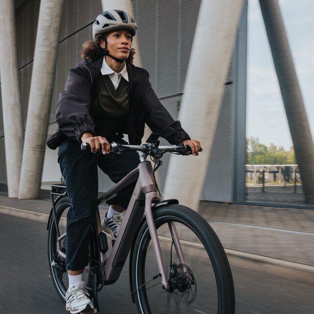 Une femme en tenue business-casual, portant un casque sur un VAE moderne, passe devant une impressionnante façade architecturale en béton et en verre. L’image saisit la dynamique de l’itinéraire urbain des trajets quotidiens.