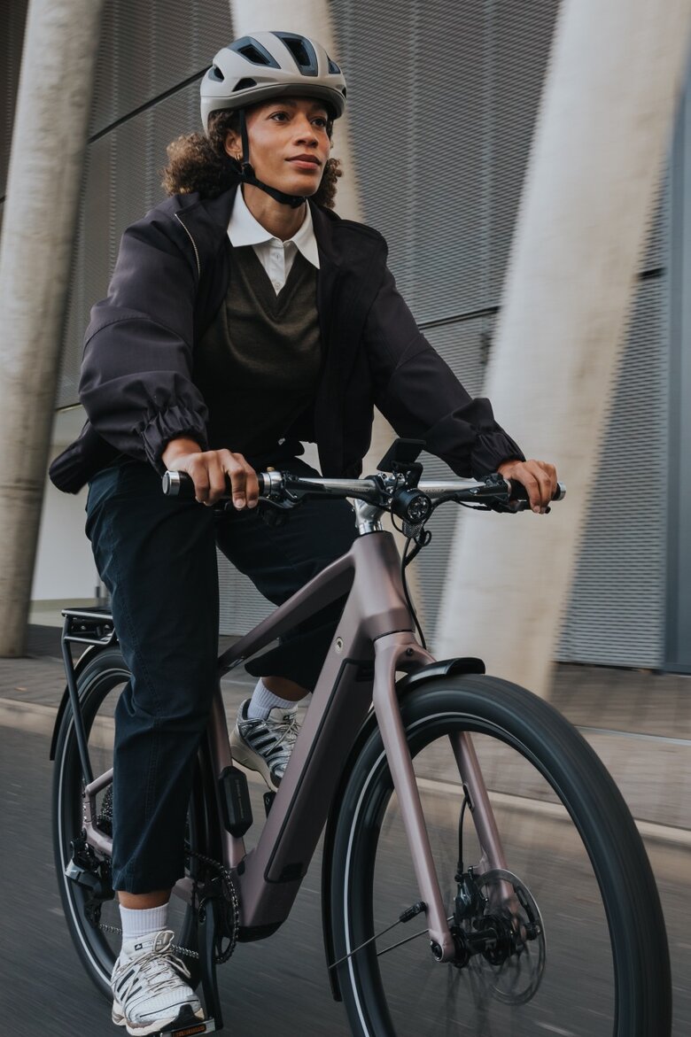 A woman in business-casual attire rides a modern eBike with a helmet past an impressive architectural façade of concrete and glass. The shot captures the dynamics of the urban commute.