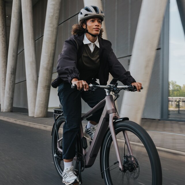 Eine Frau im Business-Casual-Look fährt mit Helm auf einem modernen eBike an einer eindrucksvollen Architekturfassade aus Beton und Glas vorbei. Die Aufnahme fängt die Dynamik der urbanen Pendlestrecke ein. 