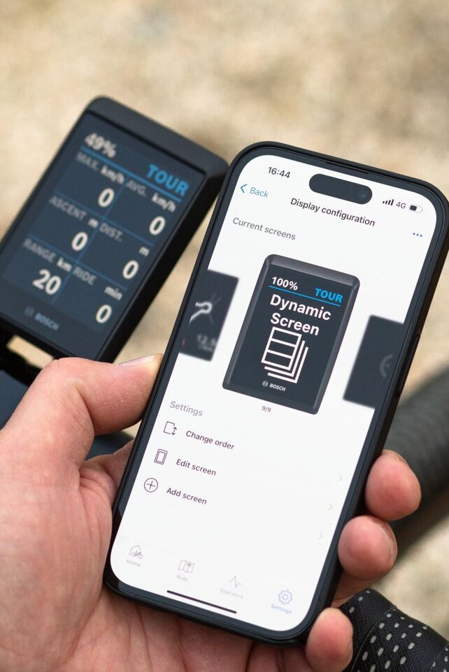 Osoba trzyma smartfon z aplikacją Bosch eBike FlowApp, na którym widoczna jest wskazanie "Konfiguracja wyświetlacza" oraz "Dynamiczny ekran". Smartfon znajduje się obok kierownicy roweru wyposażonego w cyfrowy wyświetlacz, prezentujący informacje takie jak poziom naładowania baterii, tryb (TOUR), prędkość i zasięg. Tło jest rozmyte i przedstawia otoczenie zewnętrzne.